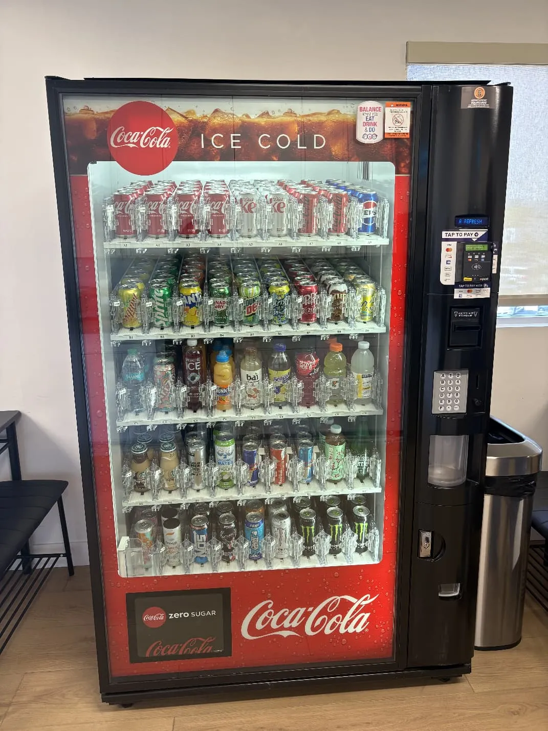 Glass-front cold drink vending machine fully stocked with sodas, juices, energy drinks and water, with tap-to-pay card reader